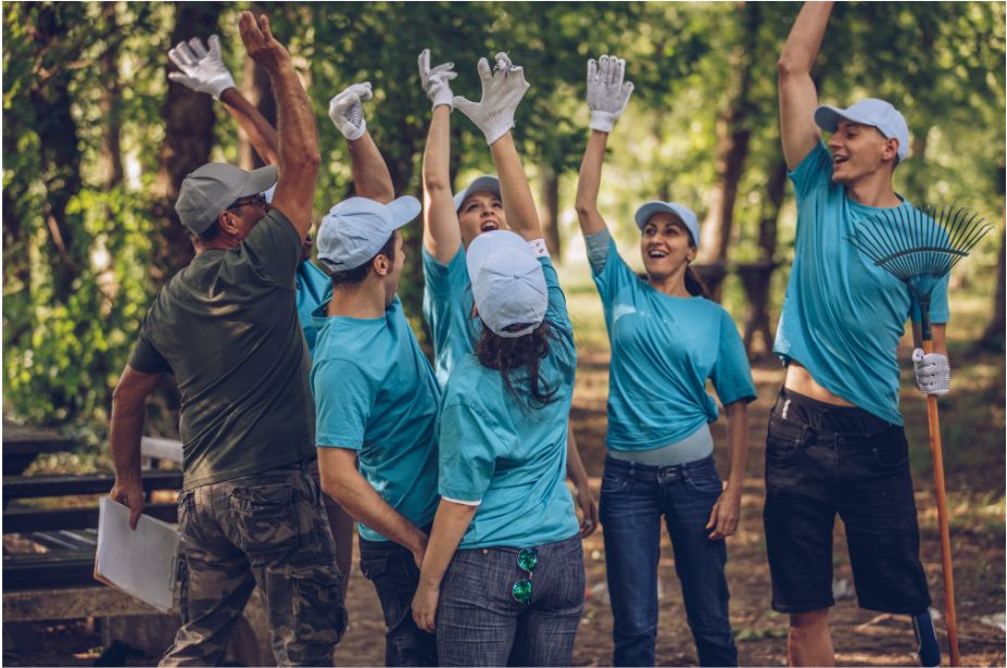 A group of volunteers in matching shirts raising their arms and cheering after finishing an outdoor clean-up.