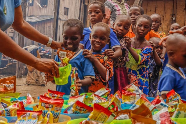 A volunteer handing packaged food to a group of children gathered around a table.