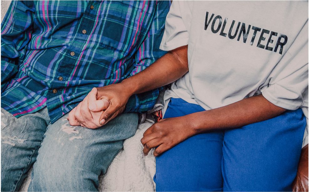 A volunteer in a Volunteer T-shirt sitting beside a community member and holding their hand.